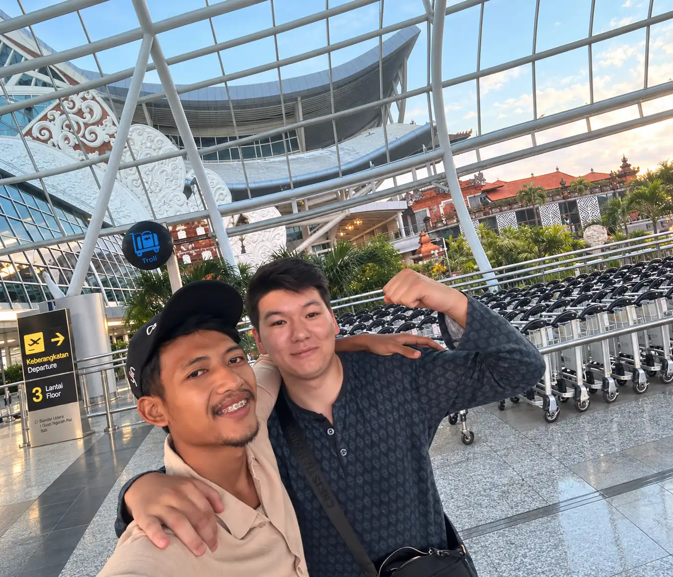 Two men taking a selfie beside luggage trolleys at Ngurah Rai Airport